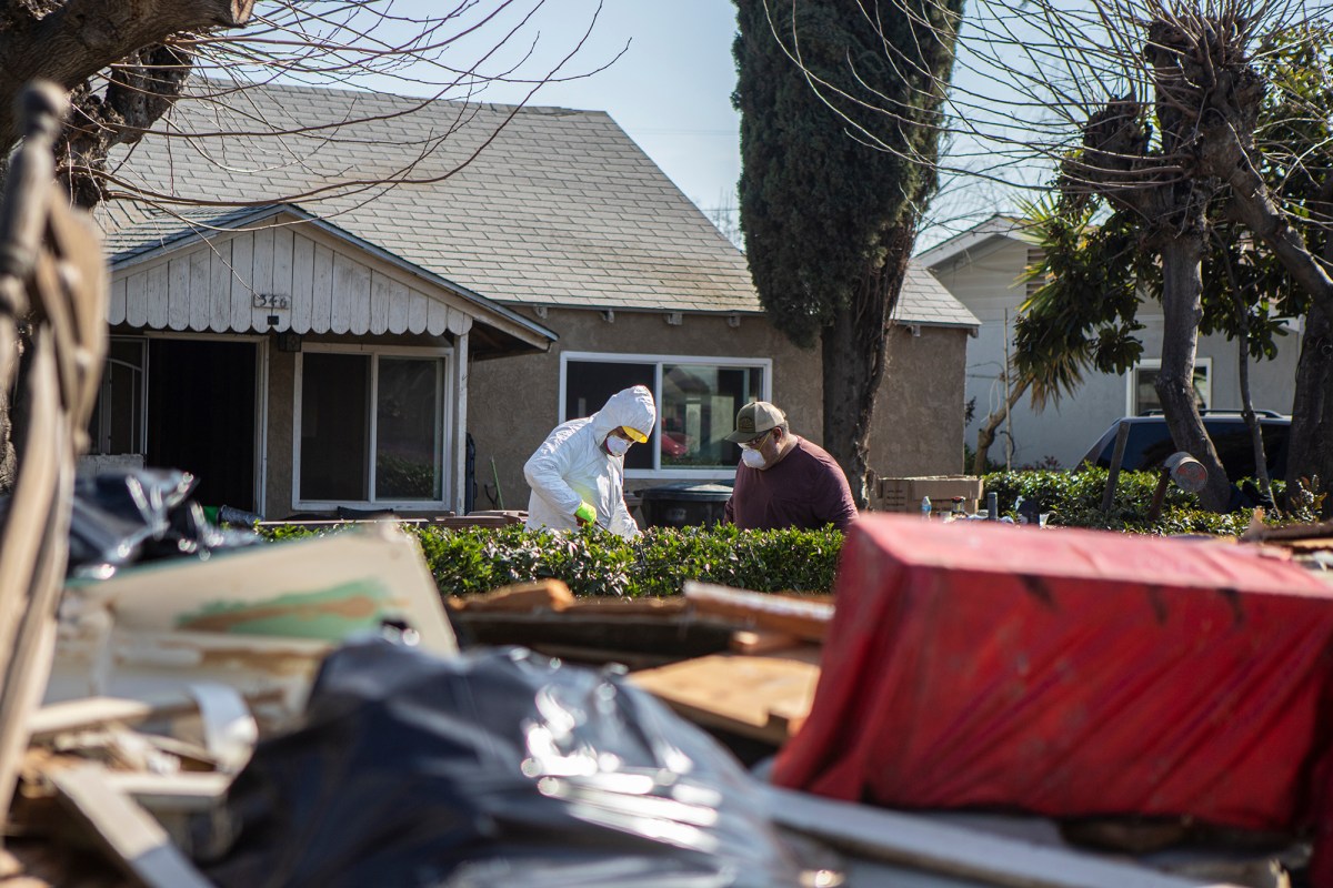 Two people in protective gear and face masks cleaning up the front year of a house with a pile of debris nearby.