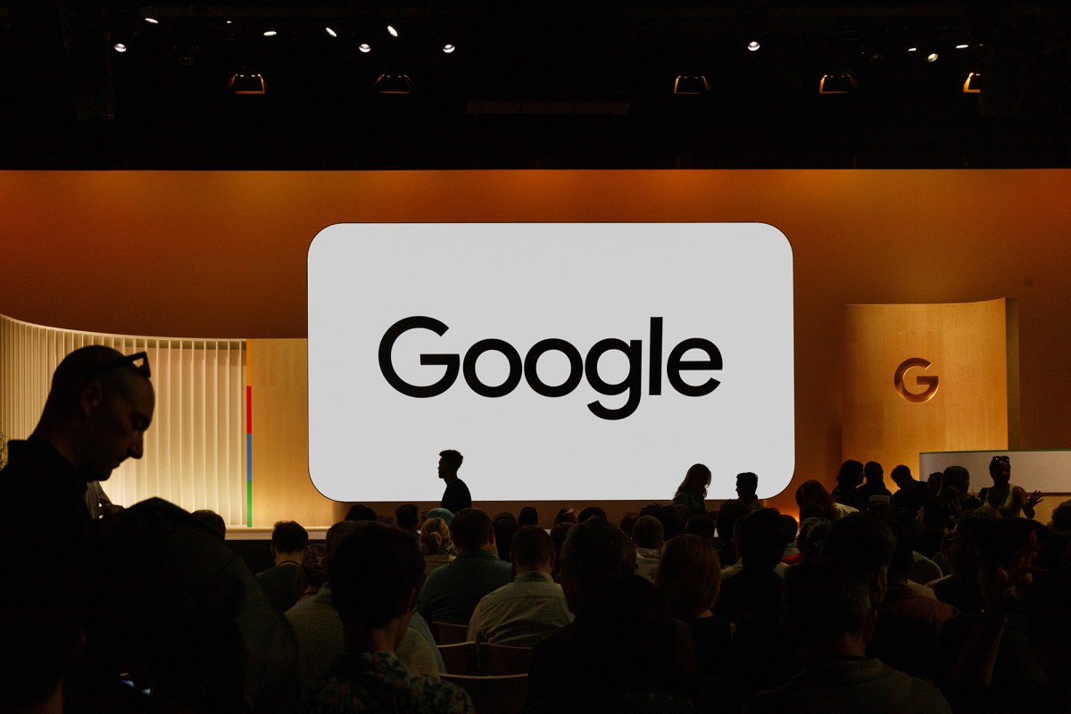 Silhouettes of people seated in a large audience, facing a stage with a prominent screen displaying the Google logo in bold black text on a white background. The stage is lit with warm lighting, creating a soft glow, while the room is dimly lit around the audience.
