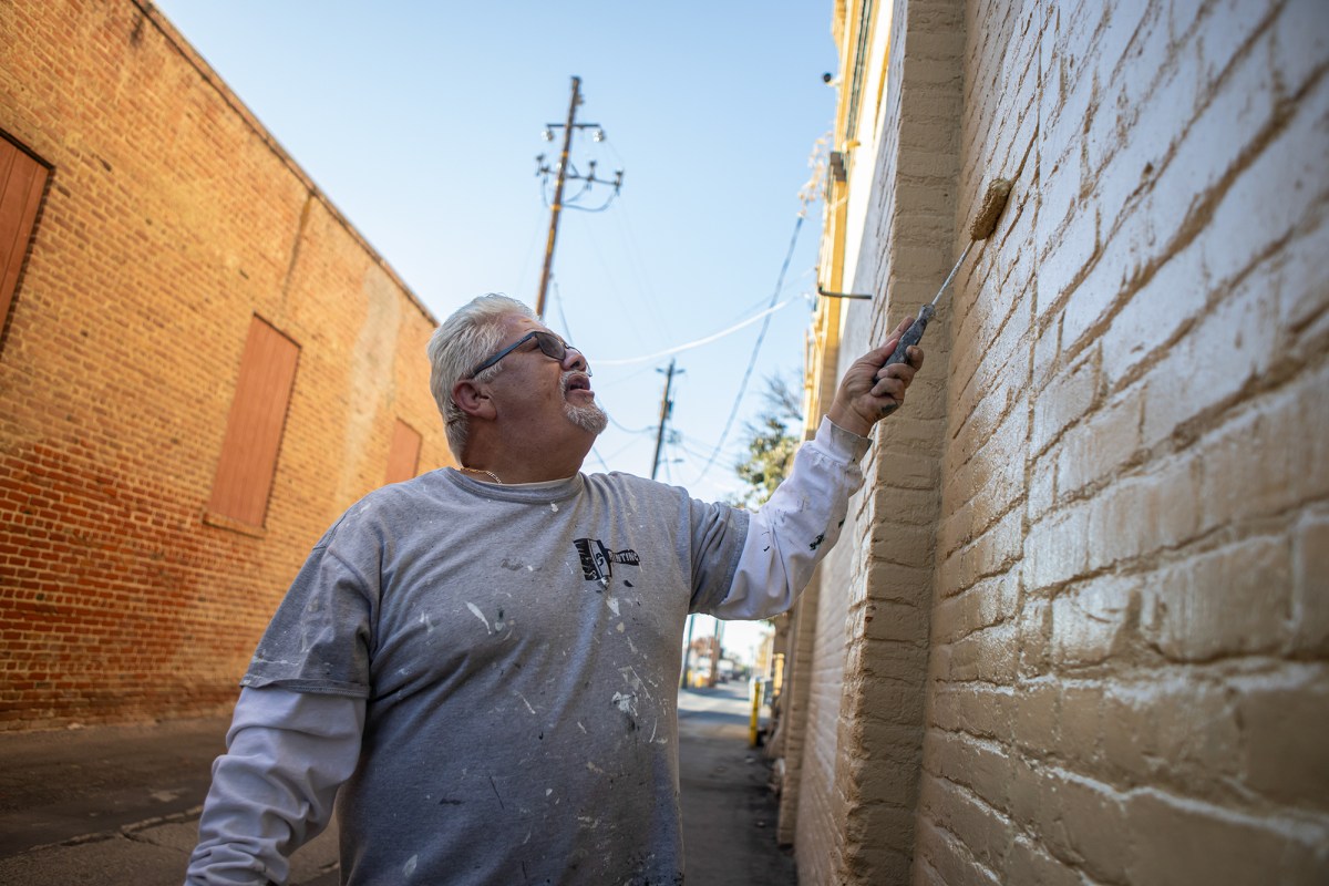 A person with white hair and wearing a grey shirt uses a paint roller to paint a yellow brick wall in an alley.