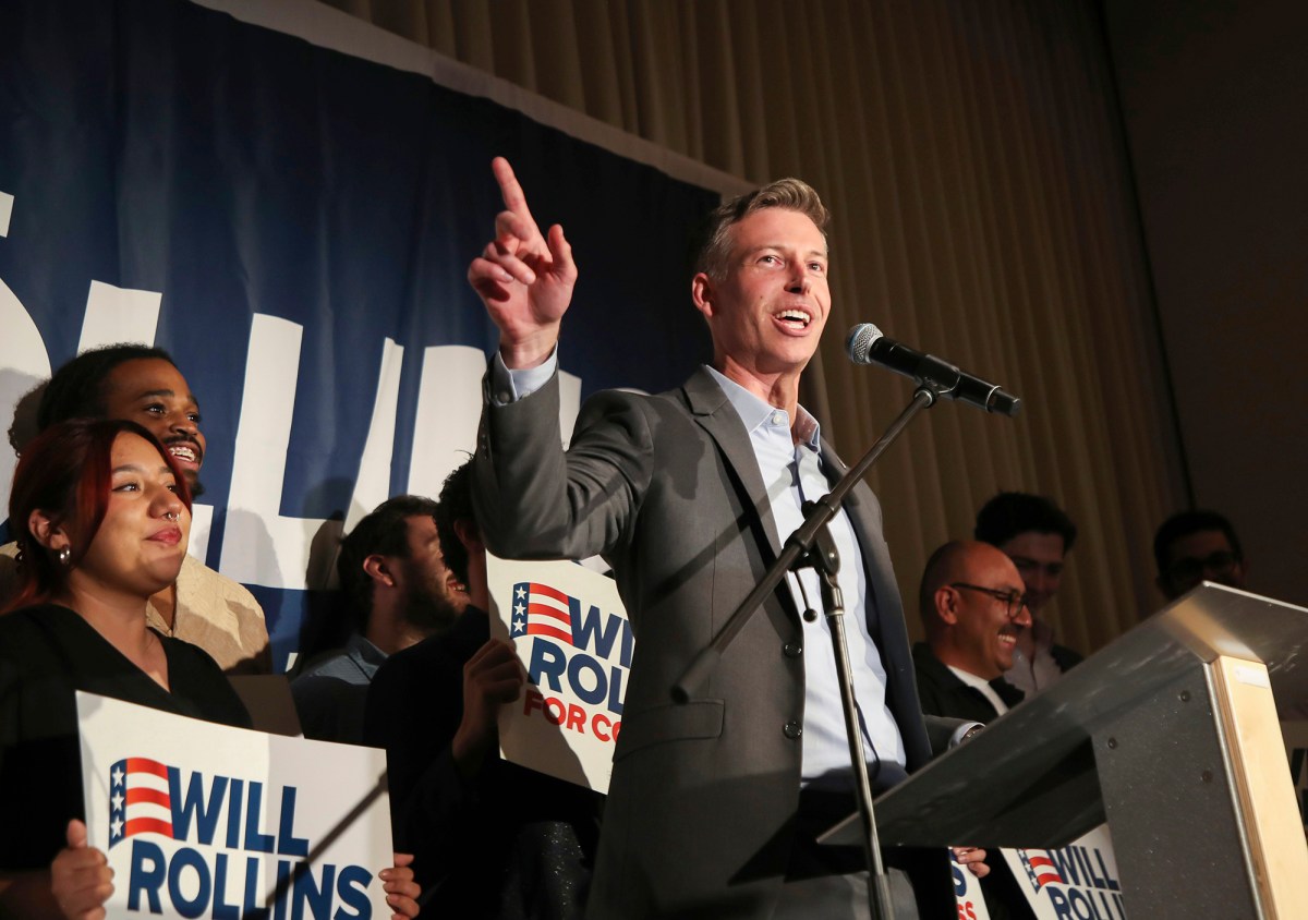 A person points as they speak at a microphone at a podium with people behind him holding up signs.