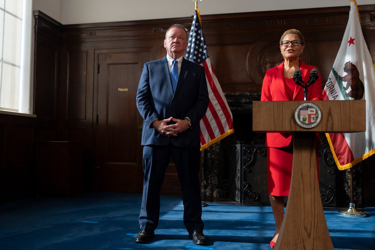 A person stands in the middle of a room with their hands folded as another person stands talking into a microphone at a podium.