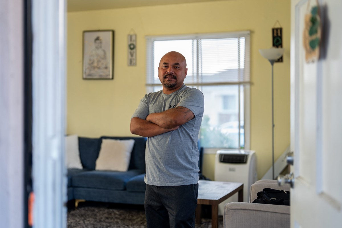A view of a man wearing a gray shirt and black pants standing in the living room of his home. A frame Buddha art piece and decorations that read "love" and "home" can be seen in the background. Chanthon Bun stands in the living room of his home in San Leandro on Nov. 23, 2024. Bun was born in Cambodia during the genocide in the 1970s and grew up in a refugee camp in Thailand before moving to the United States, where he was convicted of a crime and at age 18. That was decades ago, but now he's anxious about President-elect Donald Trump's plan for mass immigrant deportation. Photo by Jungho Kim for CalMatters